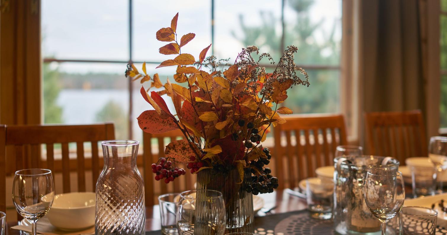 Syksyinen kukka-asetelma ruokap&ouml;yd&auml;ll&auml;, taustalla huvilan suuri ikkuna ja n&auml;kym&auml; j&auml;rvelle.