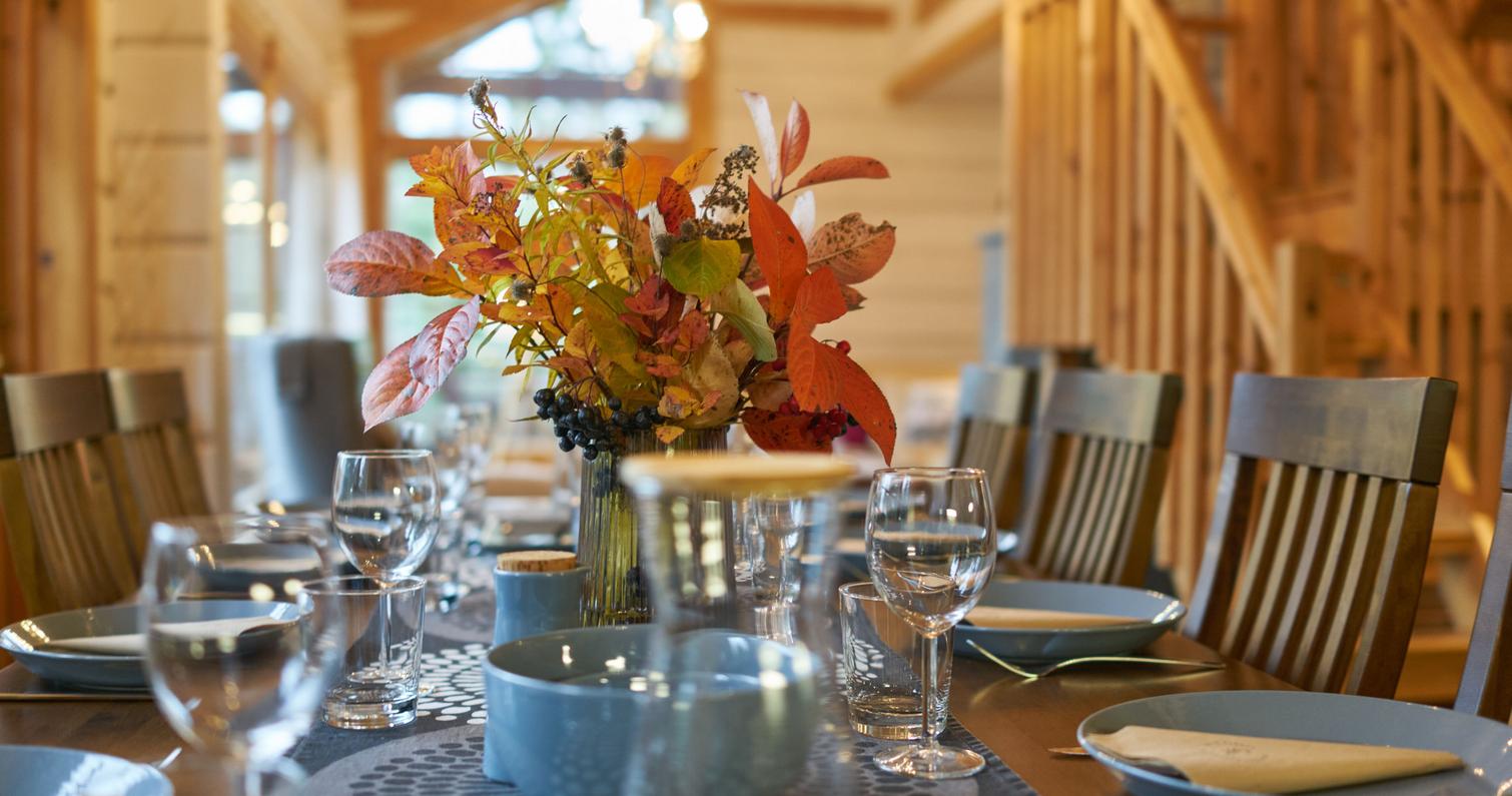Ruokap&ouml;yt&auml; kauniisti katettuna syksyisell&auml; teemalla, kukka-asetelmia ja lasiastiat, taustalla huvilan l&auml;mminhenkinen sisustus ja puuportaikko.