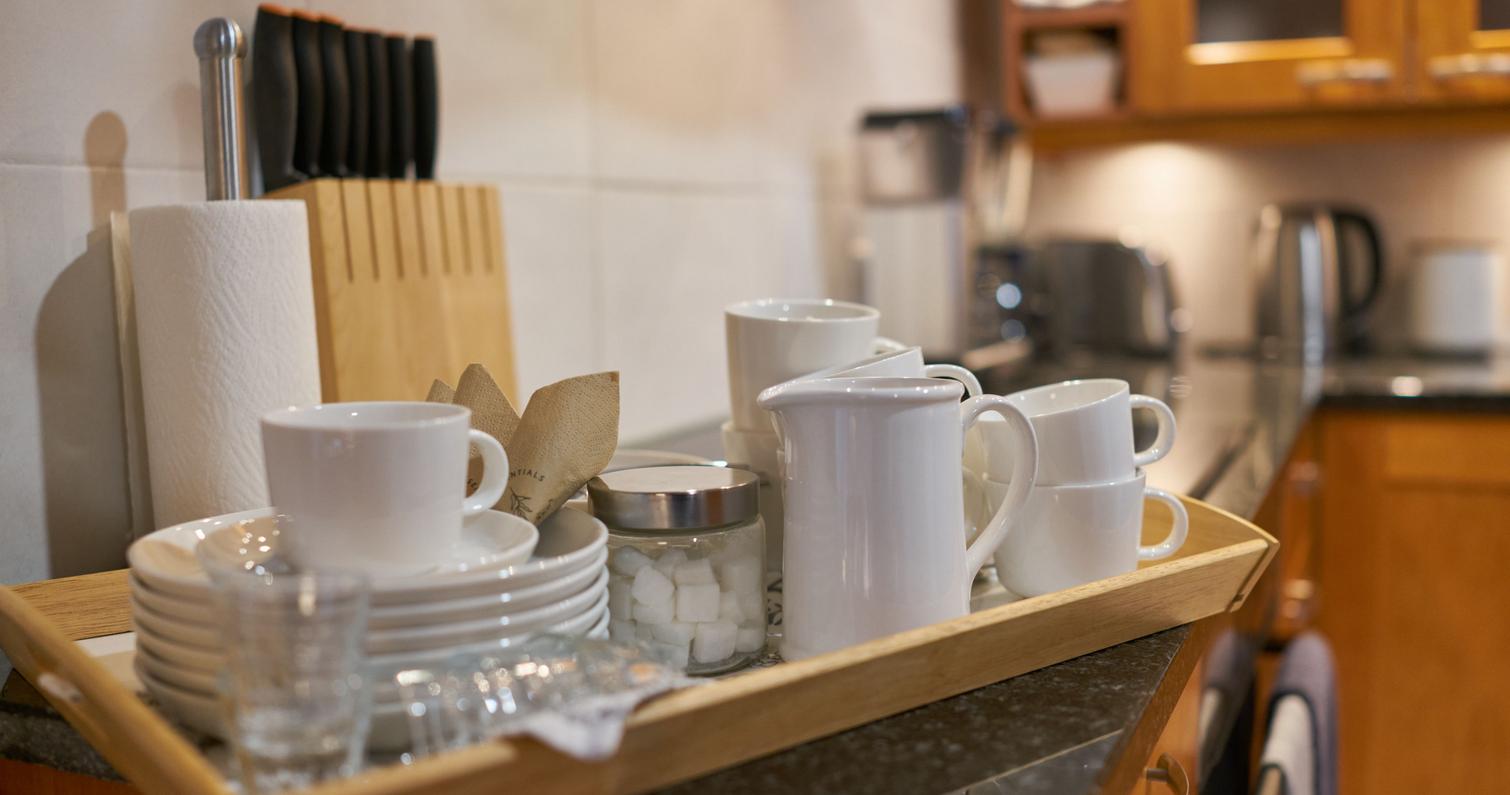 Ein Tablett mit Kaffeetassen, Tellern und einer Kaffeekanne in der Küche der Villa, im Hintergrund Holzschränke und Küchenutensilien.